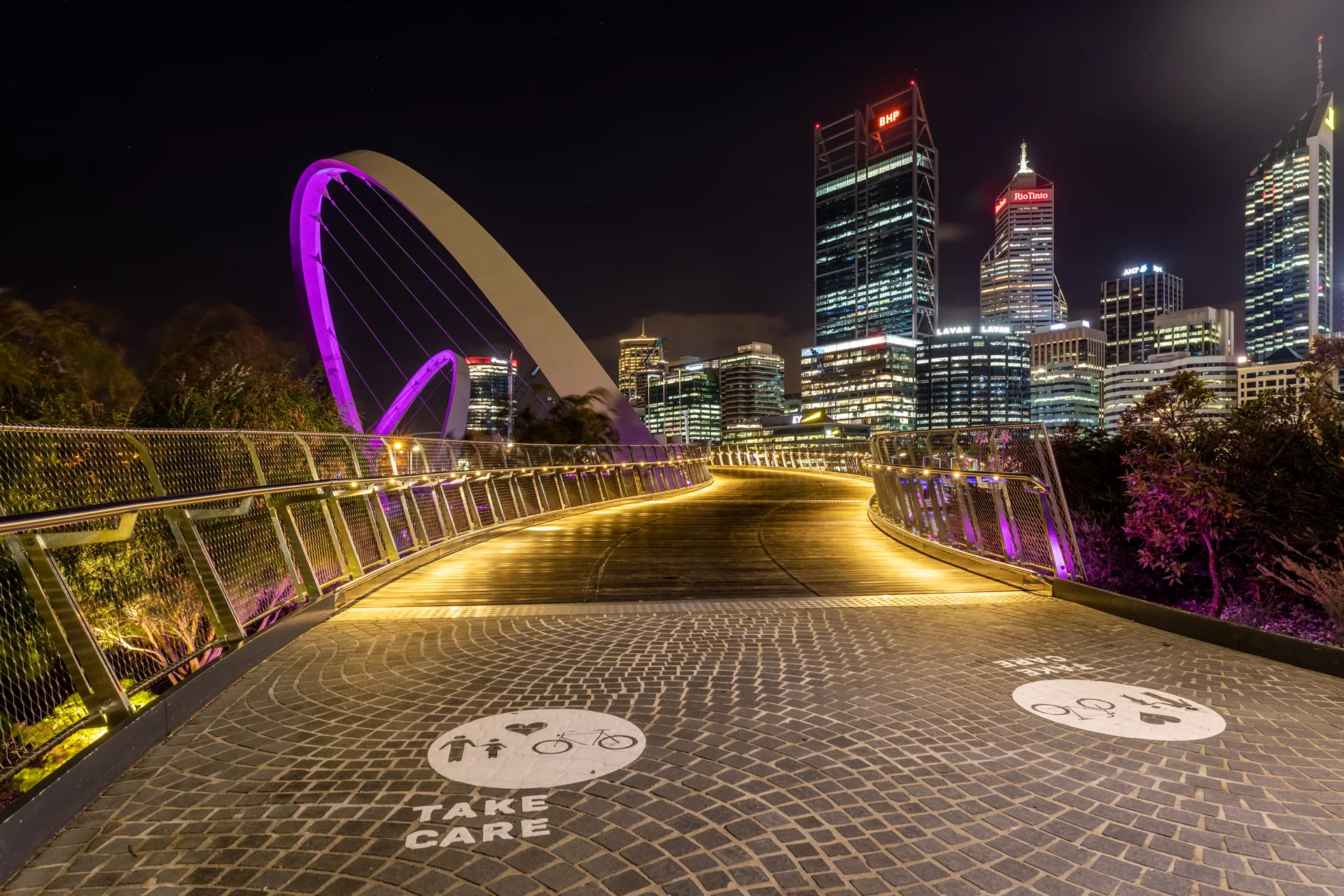 The curved Elizabeth Quay Bridge illuminated beside central Perth at night.