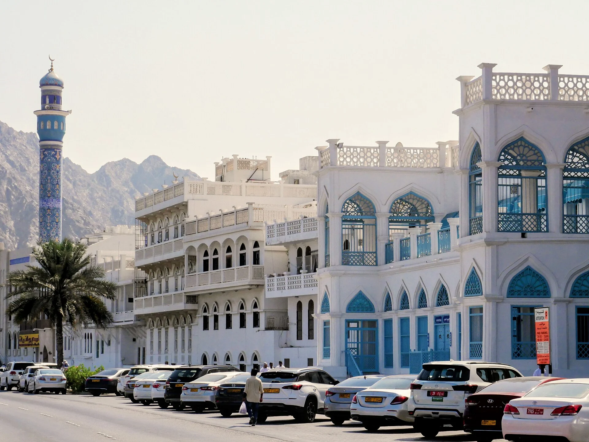 White and blue buildings along Muttrah Corniche in Muscat, with a minaret and rocky mountains behind